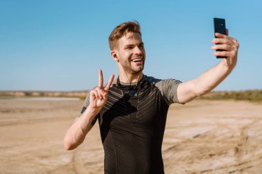 Mutlu genç sporcu sahilde dururken cep telefonuyla selfie çekiyor.