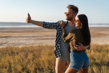 Güzel, neşeli bir çift yazın doğada dolaşırken cep telefonuyla sarılıp selfie çekiyor.