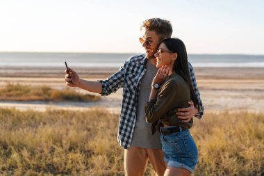 Güzel, neşeli bir çift yazın doğada dolaşırken cep telefonuyla sarılıp selfie çekiyor.