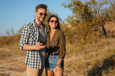 Güzel, neşeli bir çift yazın doğada dolaşırken sarılıyor ve cep telefonu kullanıyor.