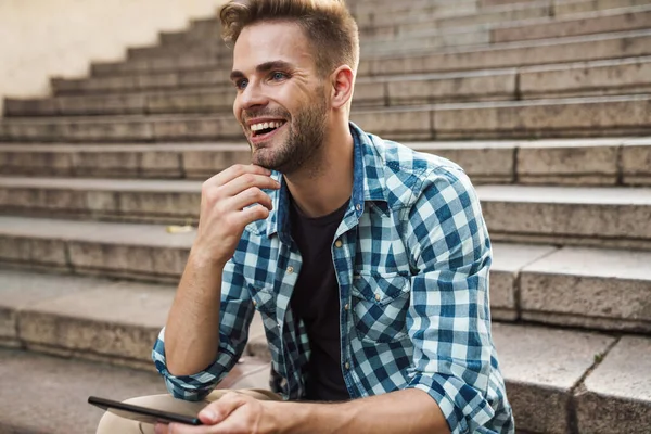 Dışarıda merdivenlerde otururken cep telefonu kullanan yakışıklı, gülümseyen genç adam.
