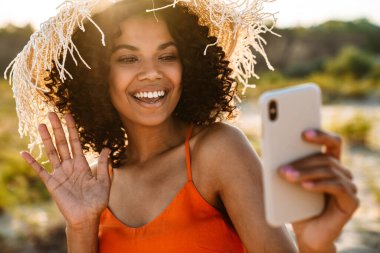 Oldukça iyimser, neşeli, Afrikalı bir kadının dışarıda sahilde cep telefonuyla görüntüsü.