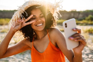 Oldukça mutlu bir Afrikalı kadının, dışarıda sahilde barış jesti yaparken cep telefonuyla selfie çekmesi.