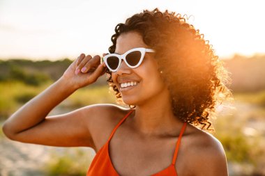 Sahilde güneş gözlükleriyle yürüyen güzel, mutlu, Afrikalı bir kadının görüntüsü.