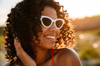 Sahilde güneş gözlükleriyle yürüyen güzel, gülümseyen Afrikalı bir kadının resmi.