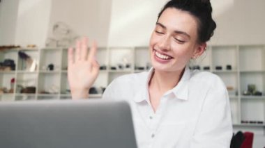 Mutlu genç bir kadın, kafede otururken bilgisayarını ve video bağlantısını kullanan biriyle konuşuyor.