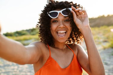 Gülümseyen Afrikalı bir kadın sahilde selfie çekiyor, güneş gözlüğü takıyor.