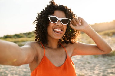 Gülümseyen Afrikalı bir kadın sahilde selfie çekiyor, güneş gözlüğü takıyor.