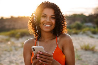 Sahilde kulaklıkla müzik dinlerken cep telefonu kullanan oldukça olumlu bir Afrikalı kadının görüntüsü