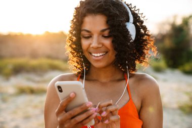 Sahilde kulaklıkla müzik dinlerken cep telefonu kullanan oldukça olumlu bir Afrikalı kadının görüntüsü