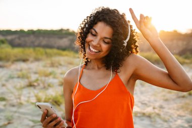 Sahilde kulaklıkla müzik dinlerken cep telefonu kullanan neşeli Afrikalı genç bir kadın.