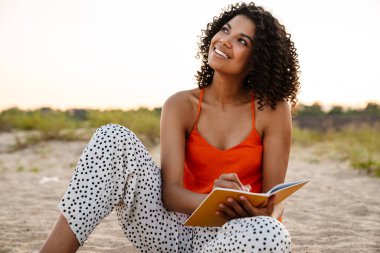 Dışarıdaki plajda not defterine notlar yazan oldukça iyimser bir Afrikalı kadın resmi.