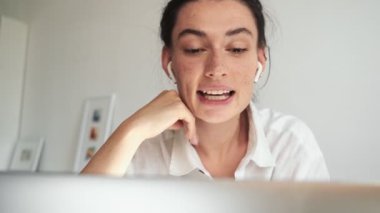 Kulaklık takan genç bir kadın odada oturmuş laptopuna bakarken biriyle konuşuyor.