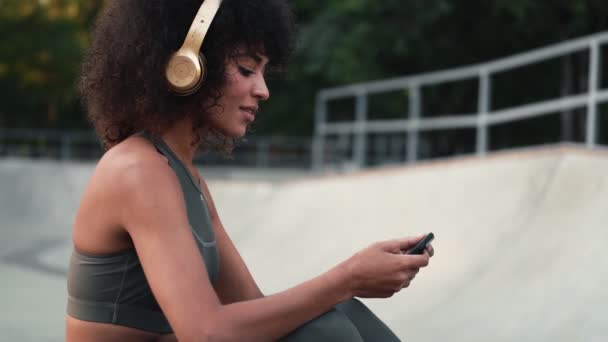 ヘッドフォンで音楽を聴きながら公園の屋外で携帯電話を使用して若いアフリカのスポーツ女性 — ストック動画