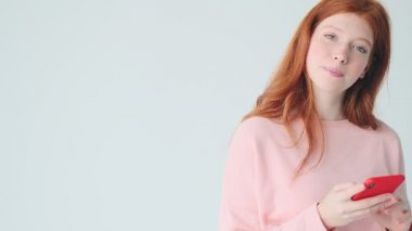 Güzel kızıl saçlı, gülümseyen kız beyaz duvar arkasında izole edilmiş cep telefonu kullanıyor.