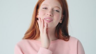 Gülen kızıl saçlı kız beyaz duvar arkasında yalnız poz veriyor.