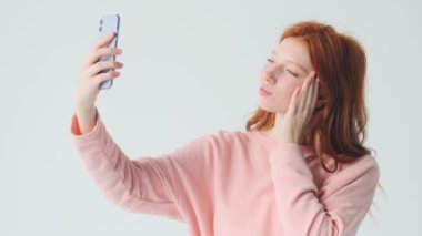 İyimser kızıl saçlı güzel kız beyaz duvar arkasında izole edilmiş cep telefonuyla selfie çekiyor.