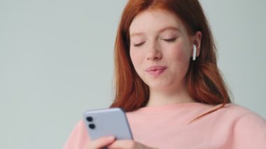 Kızıl saçlı neşeli kız, kulaklıklarıyla cep telefonu kullanıyor. Beyaz duvar arkasında izole edilmiş.