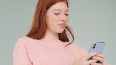 Kızıl saçlı tatlı kız, kulaklıklarıyla cep telefonu kullanıyor. Beyaz duvar arkasında izole edilmiş.