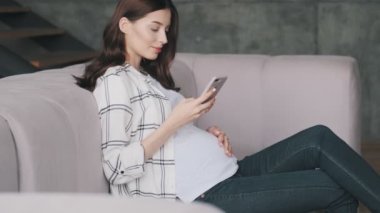 Evde cep telefonu kullanan, hamile, iyimser esmer bir kadın.