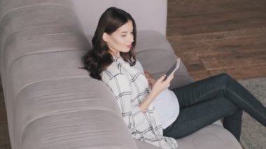 Genç hamile, mutlu, neşeli esmer kadın cep telefonunu evde kullanıyor.
