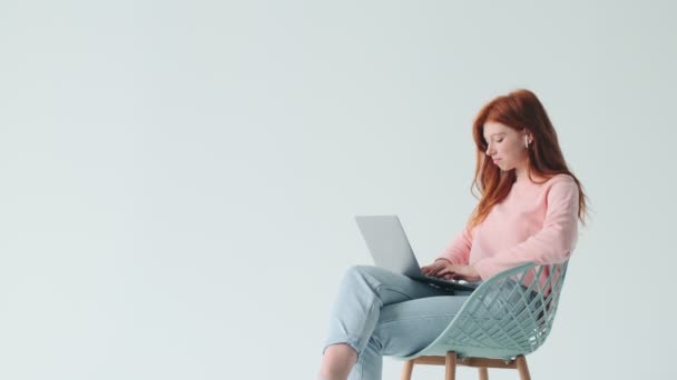 楽観的な若いです赤毛かわいい女の子使用してノートパソコンでイヤフォン白い壁の背景に隔離された — ストック動画