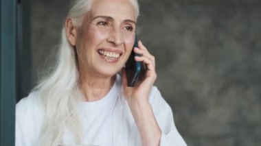 Evde cep telefonuyla konuşan ve kahve içen mutlu, kıdemli bir kadın.