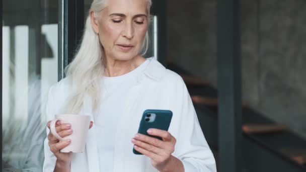 Optimistic Smiling Senior Woman Using Mobile Phone Indoors Home Drinking — Stock video