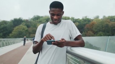Çekici bir Afrikalı Amerikalı adam akıllı telefonunu şehir köprüsünde duruyor.