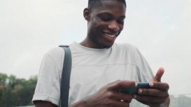 Mutlu bir Afrikalı Amerikalı adam akıllı telefonuyla şehir köprüsünde bir şey izliyor.