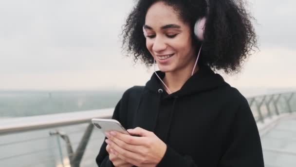 黒いパーカーを着たヘッドフォンを着た笑顔の若いアメリカ人女性が街の橋を歩いています — ストック動画