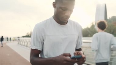 Odaklanmış bir Afrikalı Amerikalı akıllı telefonunu şehir köprüsünde yürürken kullanıyor.