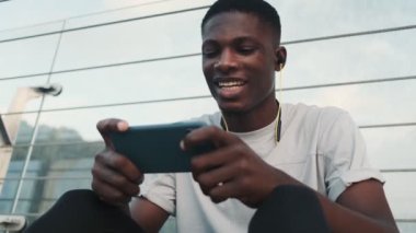 Kulaklıklı, gülümseyen Afro-Amerikalı bir adam şehir köprüsünün dışında oturan akıllı telefonuyla bir şey izliyor.