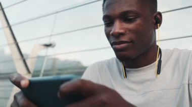 Kulaklıklı, memnun bir Afro-Amerikan adam şehir köprüsünün dışında oturan akıllı telefonuyla bir şey izliyor.