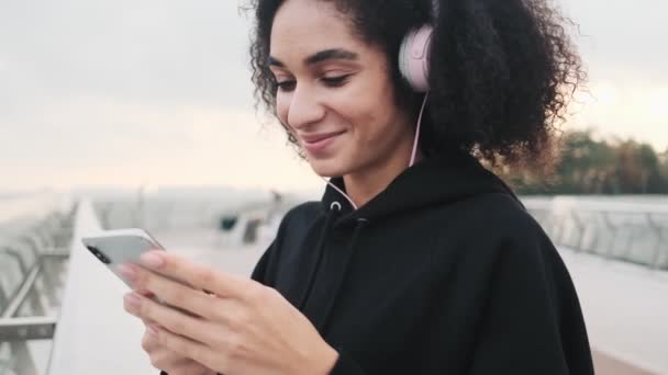 Usmívající Mladá Američanka Sluchátky Používá Svůj Smartphone Při Chůzi Městském — Stock video