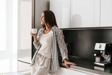 Genç, güzel, beyaz bir kadın. Gülümsüyor ve evde kahve içiyor.