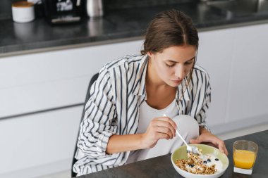 Genç beyaz kadın evde kahvaltı ederken masada oturuyor.
