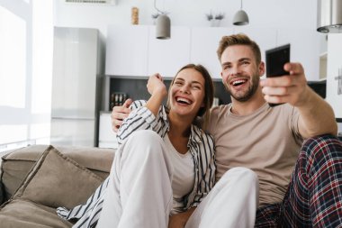 Kafkasyalı genç bir çift kanepede oturuyor ve ellerinde uzaktan kumandayla evde televizyon seyrediyor.
