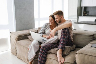 Beyaz tenli güzel bir çift evdeki kanepede oturuyor ve dizüstü bilgisayarlarını tutuyorlar.