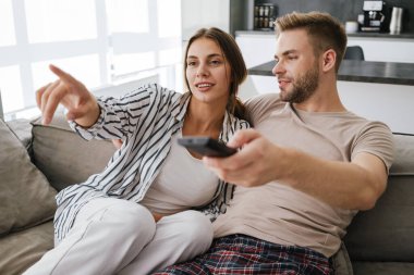 Kafkasyalı genç bir çift kanepede oturuyor ve ellerinde uzaktan kumandayla evde televizyon seyrediyor.