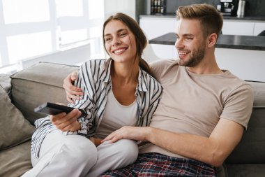 Kafkasyalı genç bir çift kanepede oturuyor ve ellerinde uzaktan kumandayla evde televizyon seyrediyor.