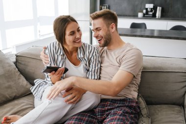 Kafkasyalı genç bir çift kanepede oturuyor ve ellerinde uzaktan kumandayla evde televizyon seyrediyor.