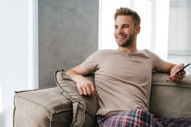 Evdeki kanepede otururken cep telefonu kullanan beyaz tenli yakışıklı bir adam.