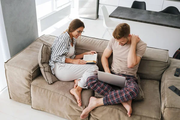 年轻的高加索夫妇坐在沙发上 手里拿着笔记本电脑 一边看书 — 图库照片