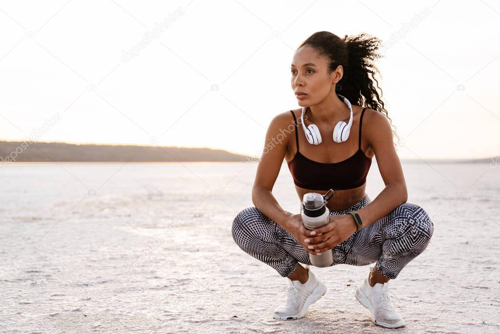 Image of young african american sportswoman squatting with water bottle on nature