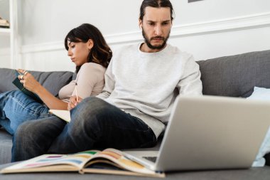 Ciddi, güzel bir çiftin kitap okuduğu ve evdeki kanepede otururken dizüstü bilgisayarla çalıştığı bir görüntü.