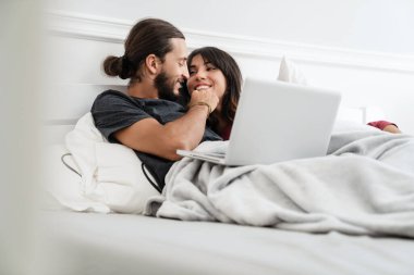 Romantik neşeli çiftin evde yatarken dizüstü bilgisayar kullanıp kucaklaşması.