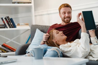 Mutlu romantik çiftin oturma odasındaki kanepede dinlenirken kucaklaşıp kitap okuduğu fotoğraf.