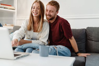 Evdeki kanepede otururken gülen ve dizüstü bilgisayar kullanan kızıl renkli bir çift.