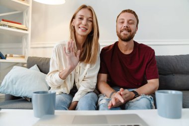 Kızıl, neşeli bir çiftin evde kanepede otururken el sallayıp dizüstü bilgisayar kullanması.